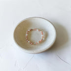 A delicate beaded bracelet with small pink and white beads rests inside a shallow, round, speckled ceramic dish on a light textured white surface.