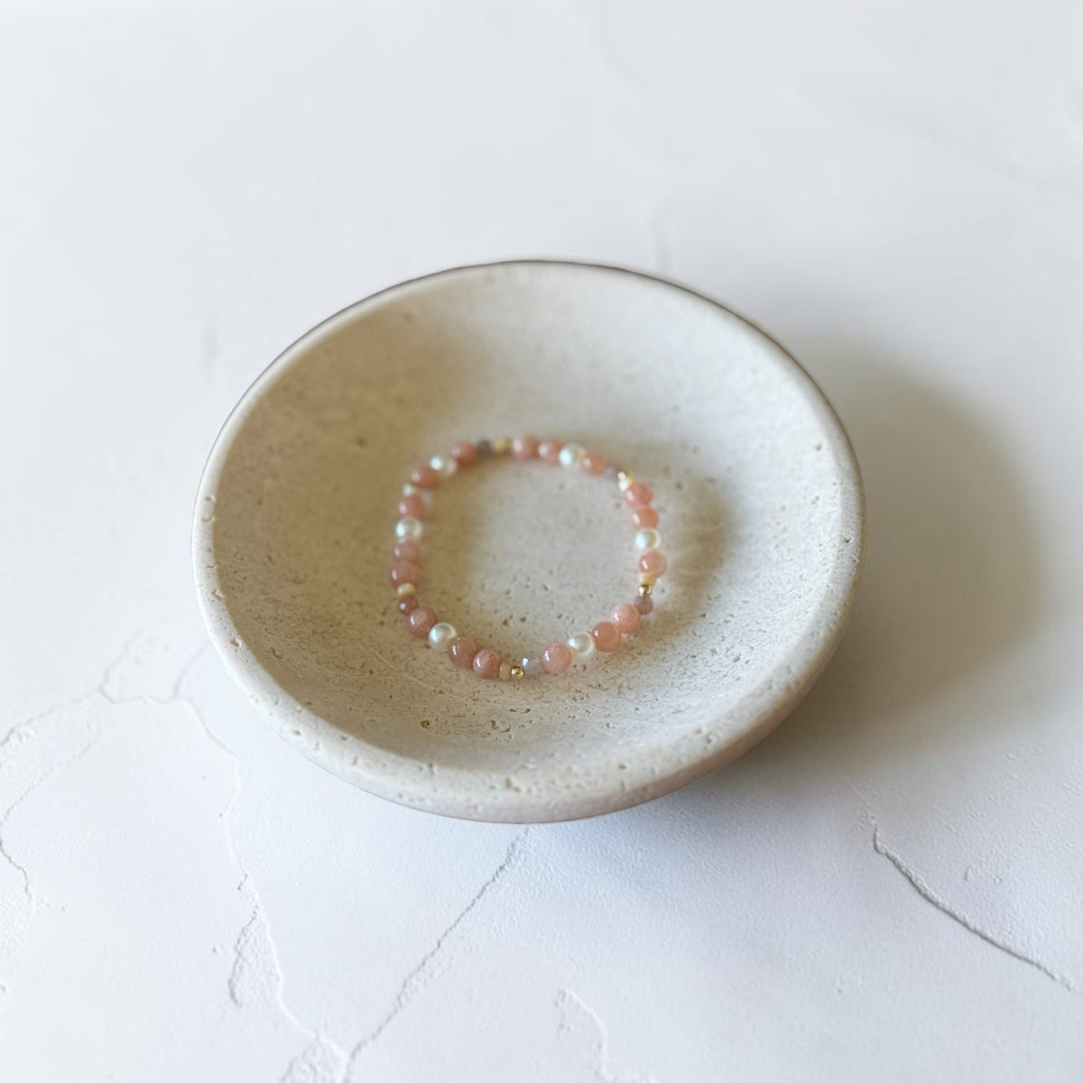 A delicate beaded bracelet with small pink and white beads rests inside a shallow, round, speckled ceramic dish on a light textured white surface.