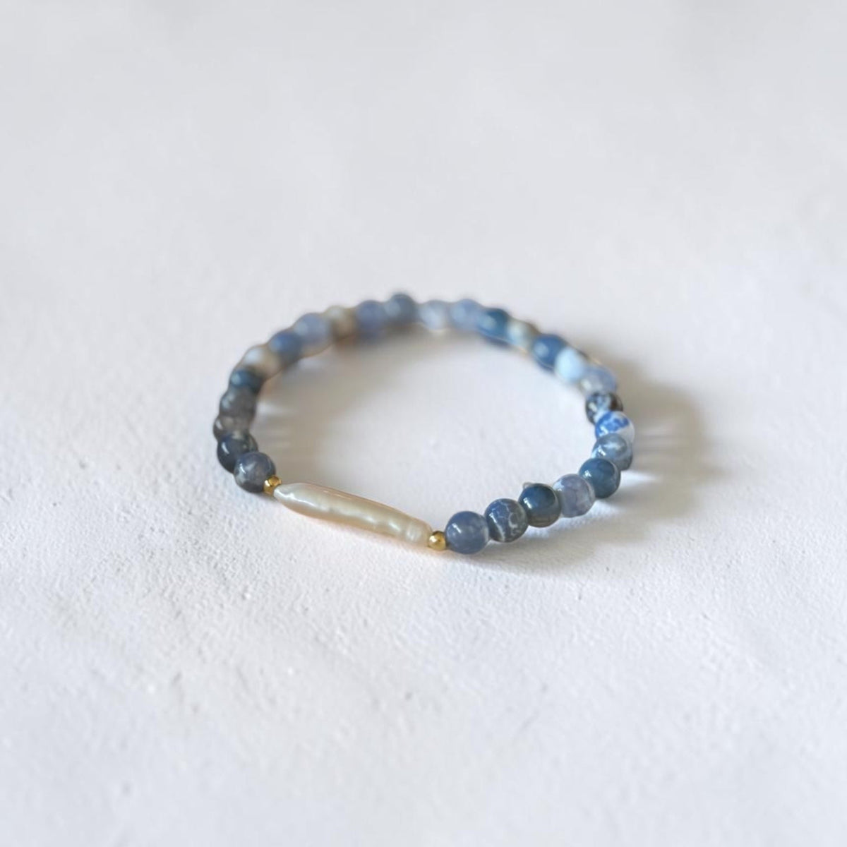 A beaded stretch bracelet featuring round blue-gray beads and a central irregular, cream-colored stone, displayed on a textured white surface.