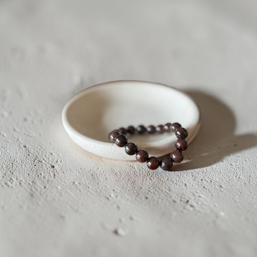 A beaded bracelet made of dark red  (brown feel) polished round beads, resting on a small white ceramic dish against a textured light gray concrete background.