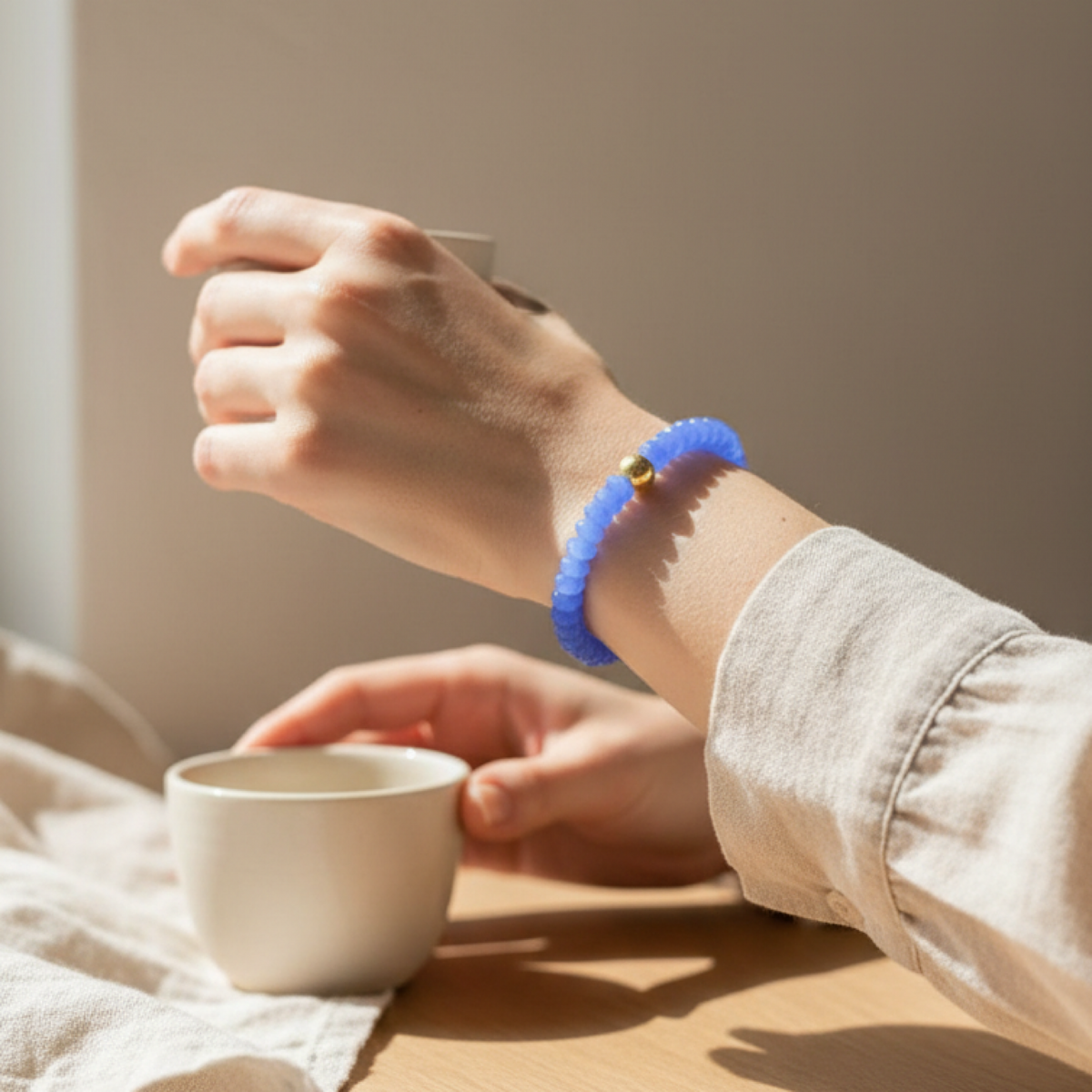 A person's wrist wearing a textured blue beaded bracelet with a small gold bead accent, shown in a warm, sunlit indoor setting with hands holding white ceramic cups.