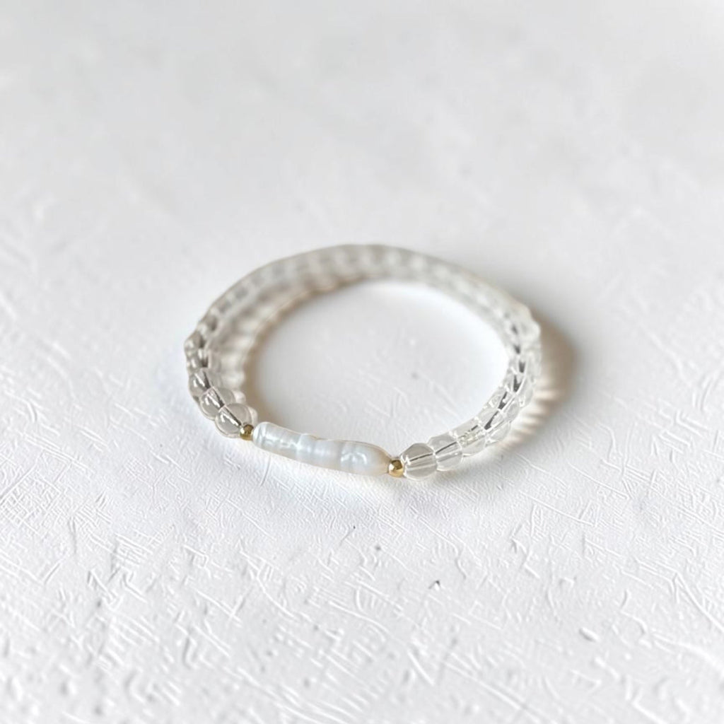 A delicate beaded stretch bracelet with clear faceted beads and a central white pearl, resting on a textured white surface.
