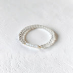 A delicate beaded stretch bracelet with clear faceted beads and a central white pearl, resting on a textured white surface.