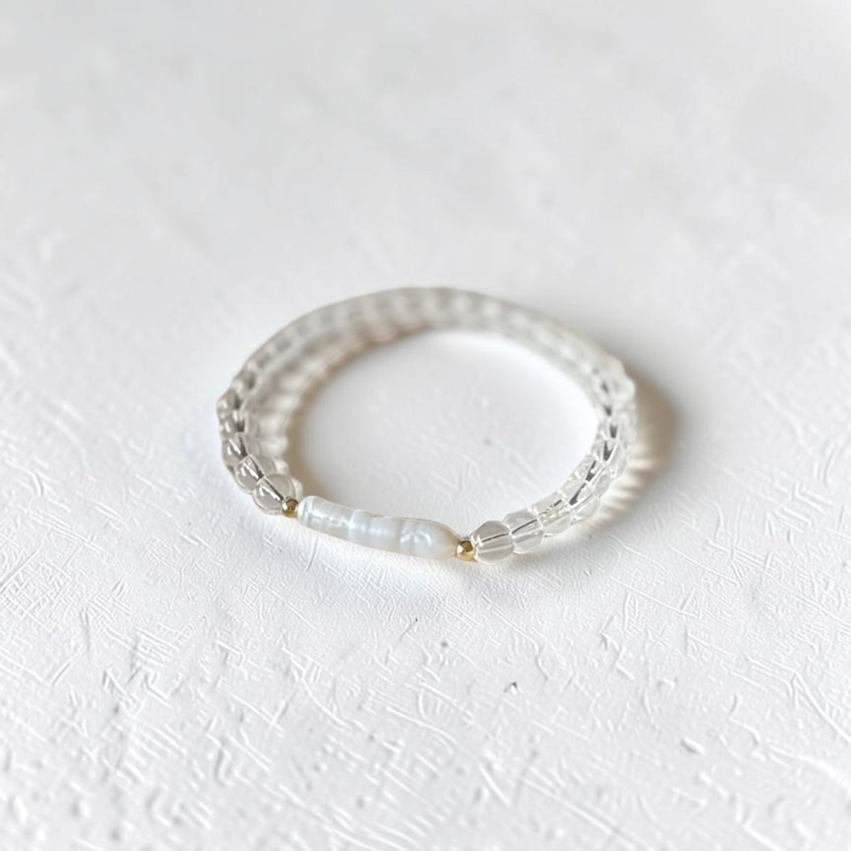 A delicate beaded stretch bracelet with clear faceted beads and a central white pearl, resting on a textured white surface.