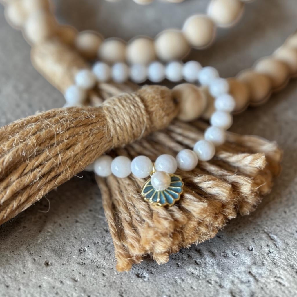 A close-up of a beaded necklace with white pearl-like round beads, a natural wood bead strand, a tan jute tassel, and a small fan-shaped enamel charm with blue and gold detailing, resting on a textured gray surface.