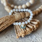A close-up of a beaded necklace with white pearl-like round beads, a natural wood bead strand, a tan jute tassel, and a small fan-shaped enamel charm with blue and gold detailing, resting on a textured gray surface.