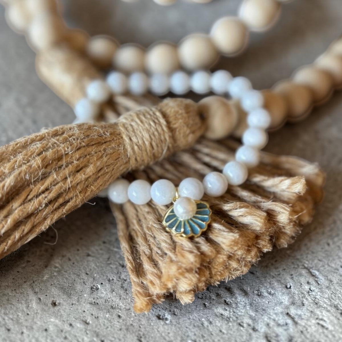A close-up of a beaded necklace with white pearl-like round beads, a natural wood bead strand, a tan jute tassel, and a small fan-shaped enamel charm with blue and gold detailing, resting on a textured gray surface.