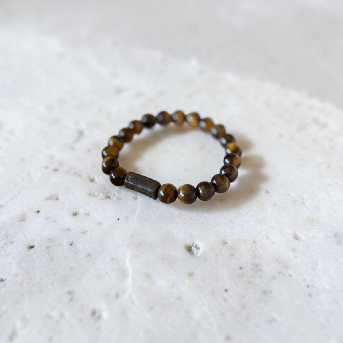 A beaded stretch bracelet made of round brown and amber tiger's eye beads with a central rectangular dark brown stone, resting on a textured white stone surface.