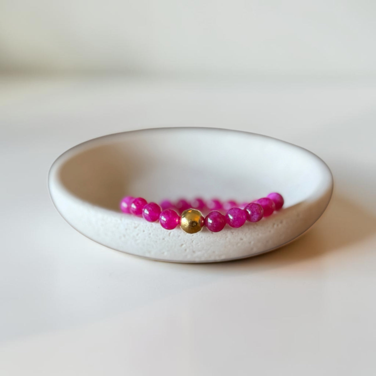 A beaded bracelet with bright pink round beads and a single central gold bead, displayed inside a small white ceramic dish on a light surface.