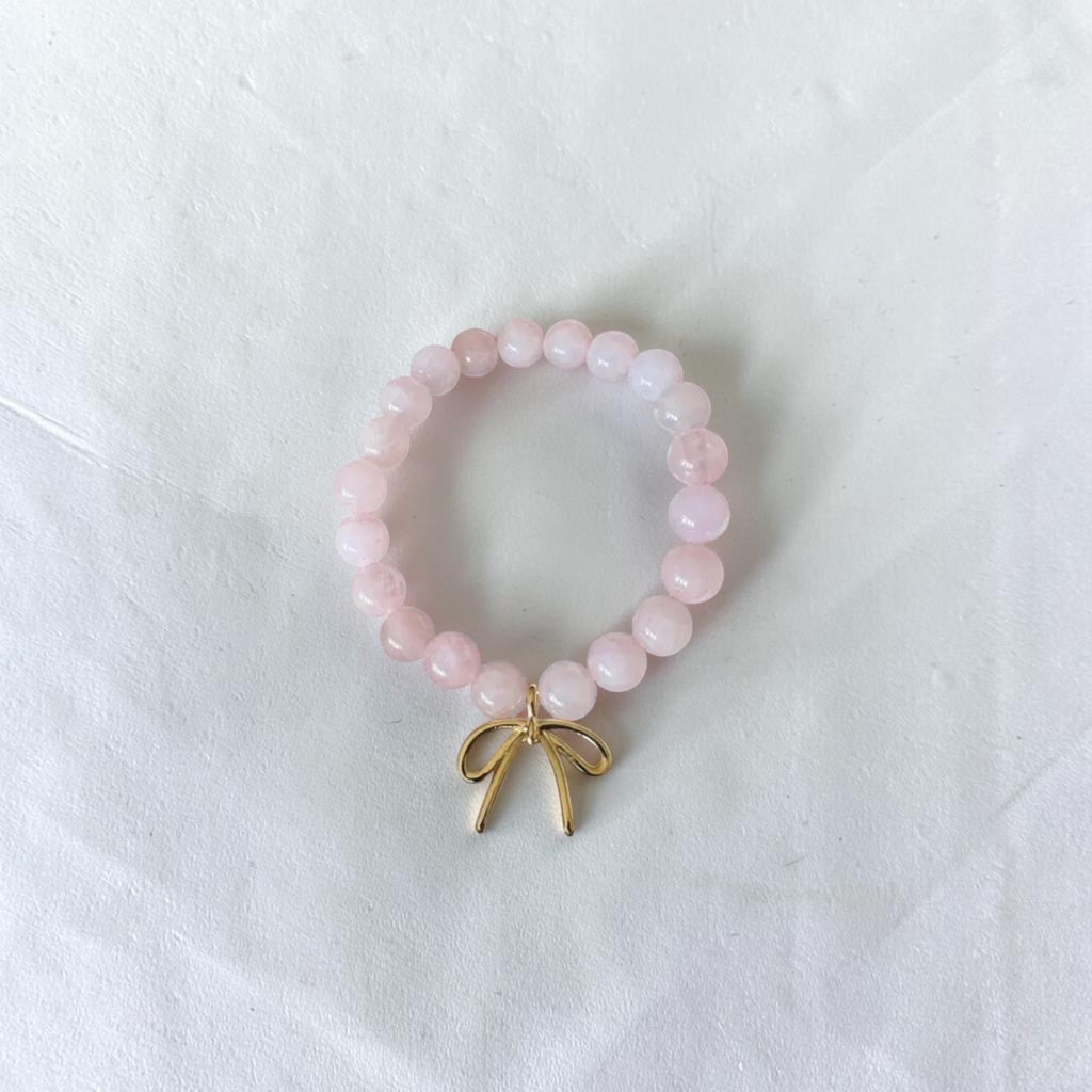 A pink beaded bracelet with a small gold bow charm, displayed on a textured white fabric background.