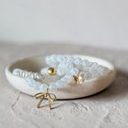 A set of delicate beaded bracelets in frosted white and clear crystal-like materials, styled in a small round ceramic dish. The set includes gold-toned accents, such as a butterfly charm, a bow charm, and a small round gold bead.