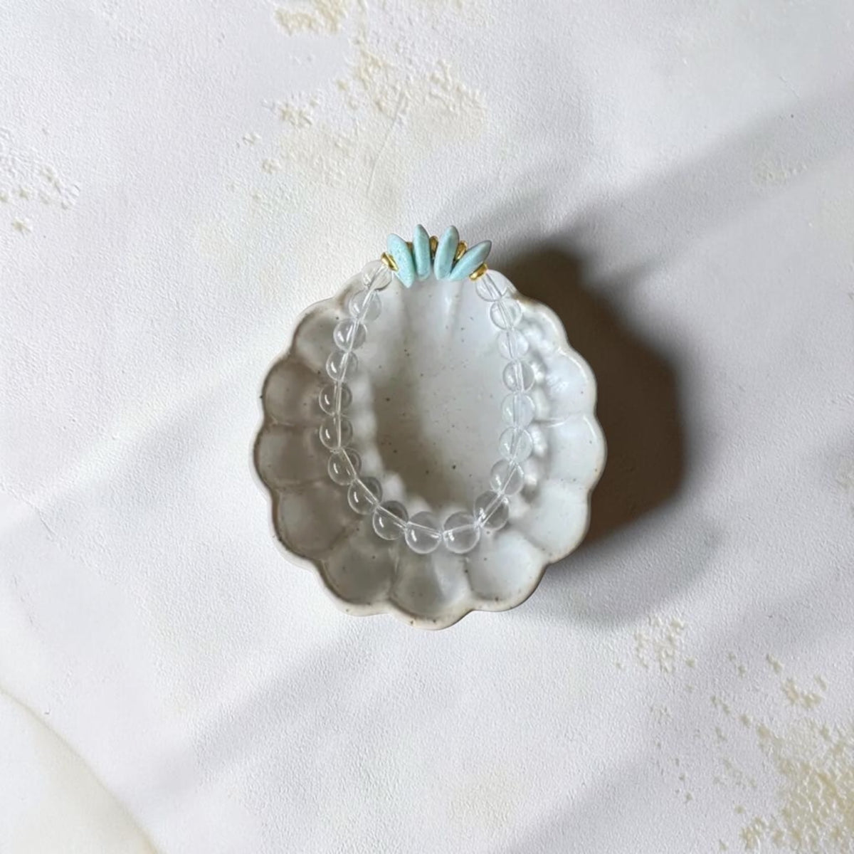A clear beaded bracelet with small light blue beads and gold-toned spacers, displayed inside a scalloped white dish on a textured off-white surface.