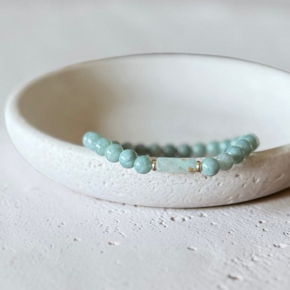 A beaded stretch bracelet in light aqua blue-green beads with a central rectangular bead and small gold spacer rings, displayed on a white ceramic dish against a textured light gray background.