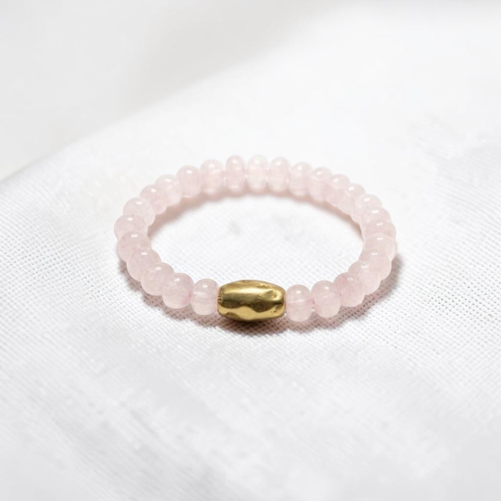 A beaded stretch bracelet made of light pink rose quartz beads with a central gold-colored metallic bead, resting on a white textured fabric background.