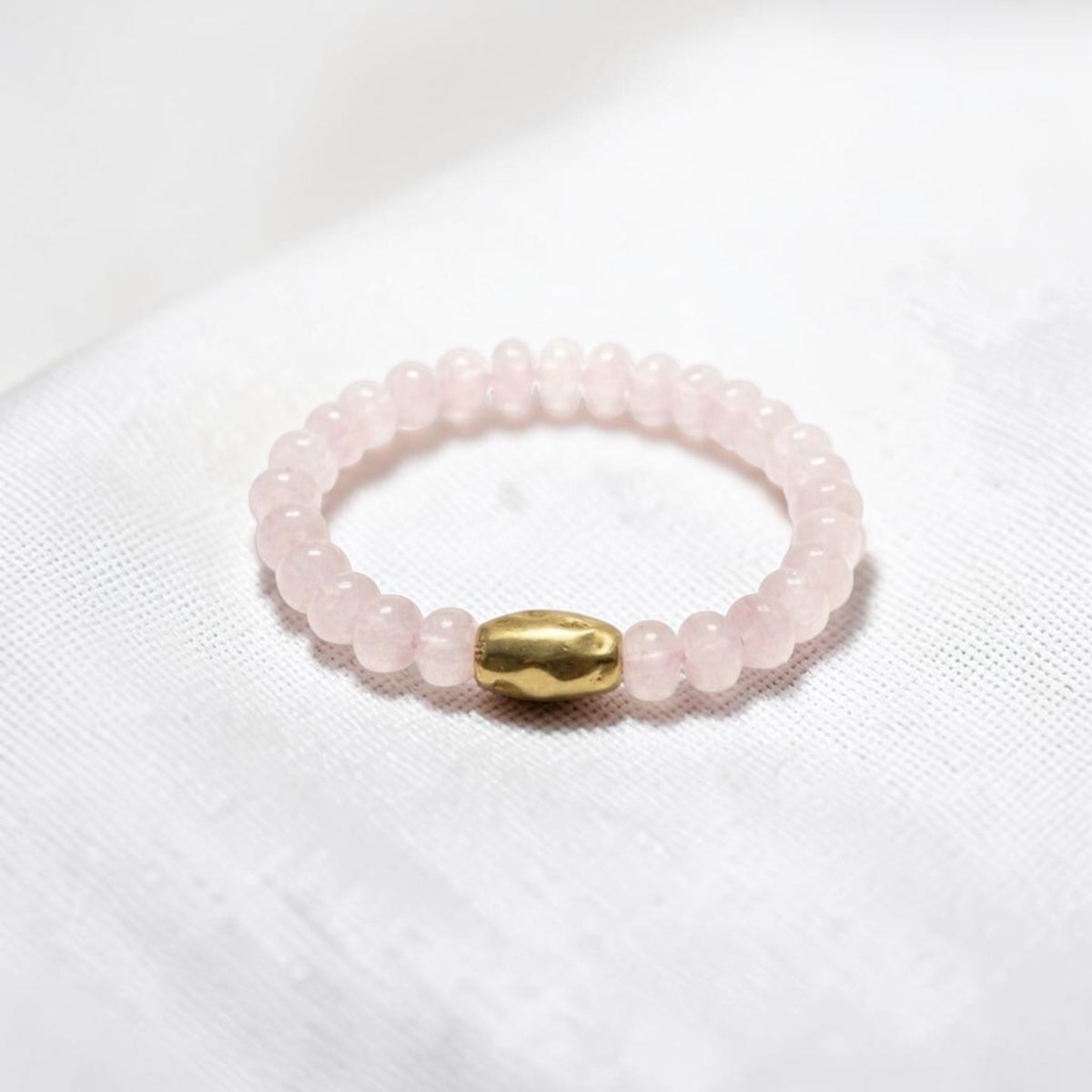 A beaded stretch bracelet made of light pink rose quartz beads with a central gold-colored metallic bead, resting on a white textured fabric background.