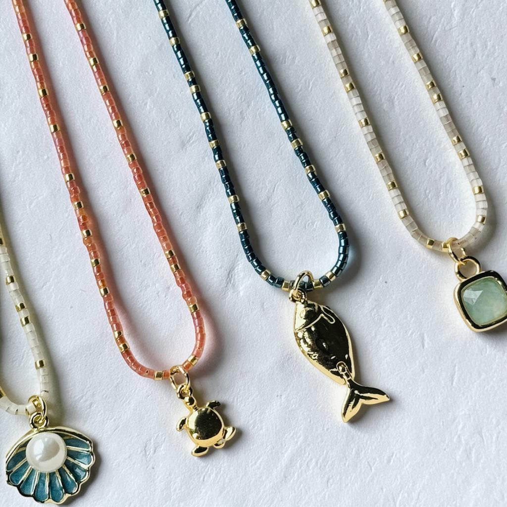 Four delicate beaded necklaces with gold-tone pendants are arranged diagonally on a textured white surface. The necklaces feature coral pink, metallic blue, and cream beads, with pendants including a gold fish, a gold turtle, a blue and white seashell with a pearl, and a light green stone.