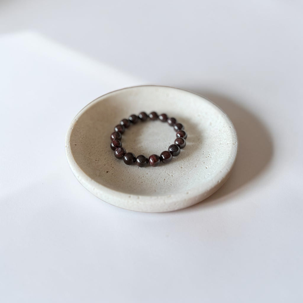 A dark brown beaded stretch bracelet rests on a small, round, textured off-white ceramic dish against a clean white background.