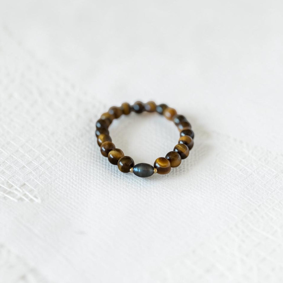 A beaded stretch bracelet featuring round brown tiger's eye beads and one smooth, dark gray oval accent bead, displayed on a textured white fabric background.
