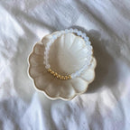 A beaded stretch bracelet with translucent white beads and a small gold section, displayed on a small scalloped ceramic dish over a white fabric background.