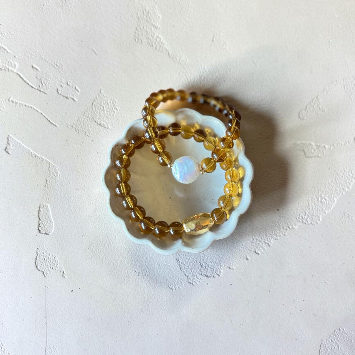 A beaded bracelet made of translucent amber-yellow beads with a small white iridescent opal-like bead, displayed on a small white scalloped dish against a textured off-white background.