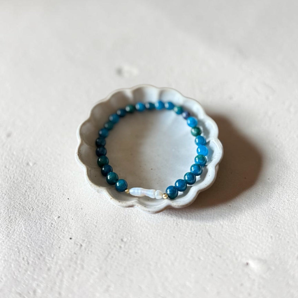 A beaded stretch bracelet with blue and teal beads and a small white accent bead, displayed on a small white scalloped dish against a textured off-white background.