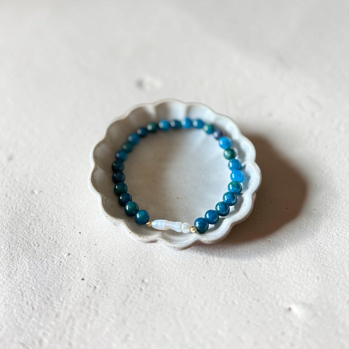 A beaded stretch bracelet with blue and teal beads and a small white accent bead, displayed on a small white scalloped dish against a textured off-white background.