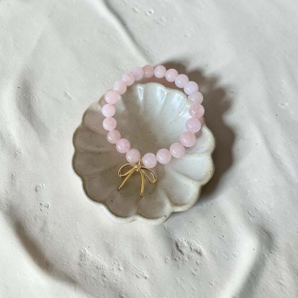 A pink beaded bracelet with a small gold bow charm, displayed on a scalloped white dish over a textured light gray surface.
