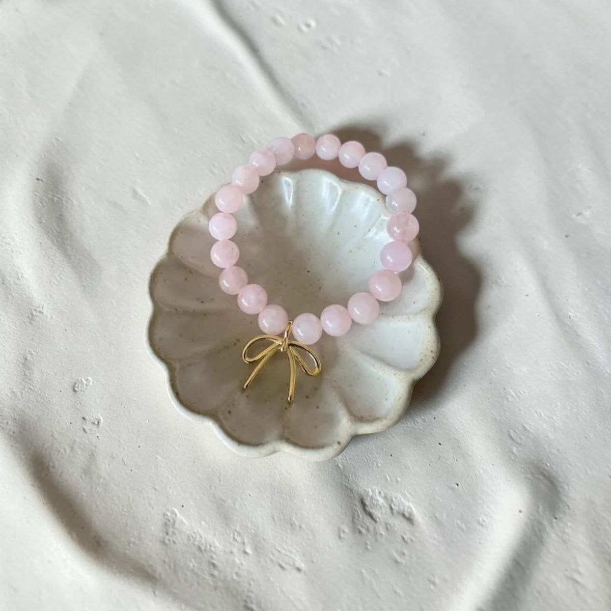 A pink beaded bracelet with a small gold bow charm, displayed on a scalloped white dish over a textured light gray surface.