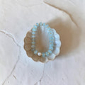 A beaded bracelet with light blue faceted beads and small gold spacer rings, displayed inside a scalloped ceramic dish on a textured off-white surface.