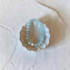 A beaded bracelet with light blue faceted beads and small gold spacer rings, displayed inside a scalloped ceramic dish on a textured off-white surface.