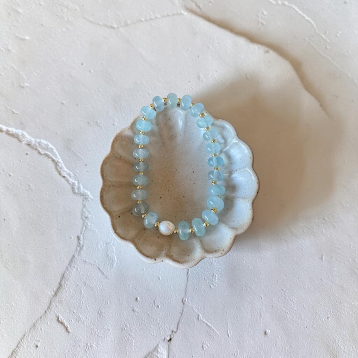 A beaded bracelet with light blue faceted beads and small gold spacer rings, displayed inside a scalloped ceramic dish on a textured off-white surface.