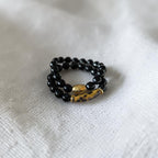 A double-strand beaded bracelet featuring glossy black beads and a central gold-toned spacer bead with a brown tiger-eye pattern, resting on a textured white fabric background.