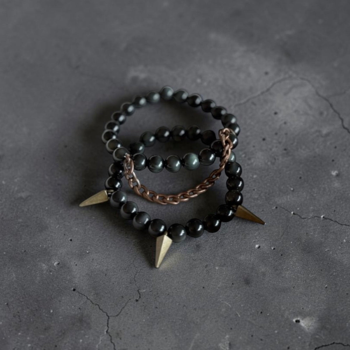 Two beaded bracelets with black beads and gold-toned spikes, one with a rose gold chain link, displayed on a dark textured surface.