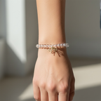 A clear beaded bracelet with a small gold bow charm, shown worn on a person's wrist against a softly lit neutral background.