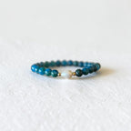 A beaded bracelet featuring dark teal blue beads and a small gold-toned accent bead, resting on a textured white surface.