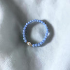 A light blue beaded bracelet with a small white teardrop-shaped bead and gold-toned accents, resting on a textured white fabric background.
