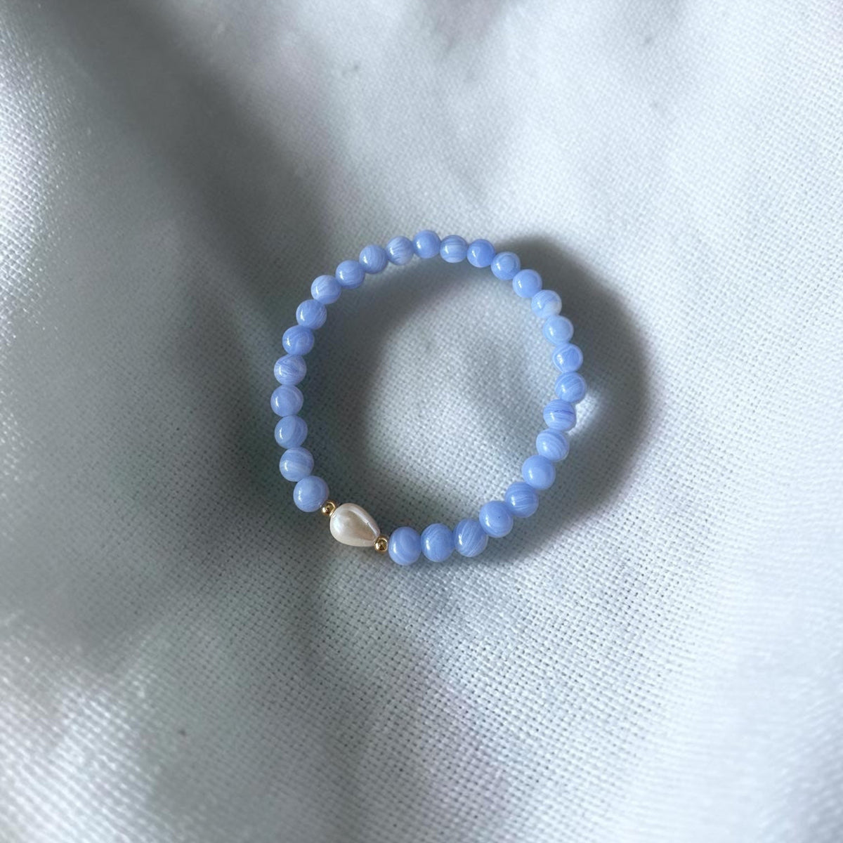 A light blue beaded bracelet with a small white teardrop-shaped bead and gold-toned accents, resting on a textured white fabric background.