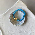 A beaded stretch bracelet with bright turquoise-blue round beads and a small gold accent bead, displayed on a scalloped white ceramic dish.