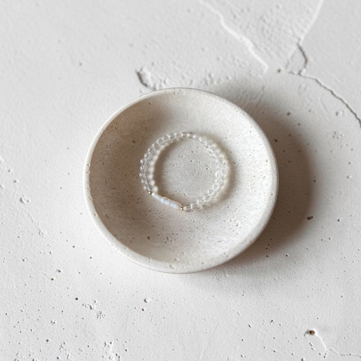 A small round ceramic dish with a speckled, matte off-white finish, holding a delicate beaded ring, set against a textured concrete background.