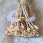A light pink beaded bracelet with a textured gold clasp, displayed on a rustic brown woven tassel against a soft beige fabric background.