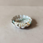 A set of delicate beaded stretch bracelets, including white pearl-like beads, light blue beads, and small brown accents, displayed on a small white ceramic dish against a neutral beige background.