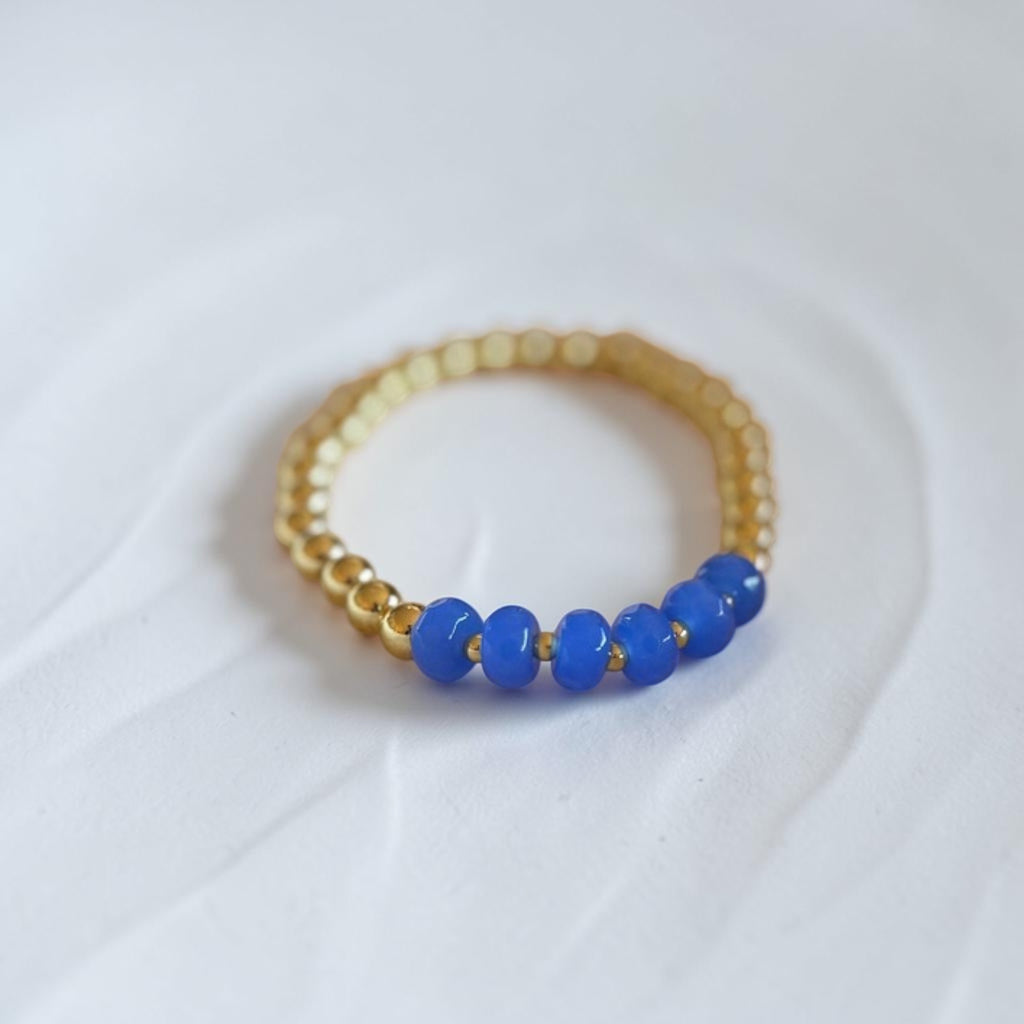 A gold beaded bracelet with five smooth blue beads on one side, photographed on a soft white fabric background.