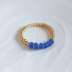 A gold beaded bracelet with five smooth blue beads on one side, photographed on a soft white fabric background.