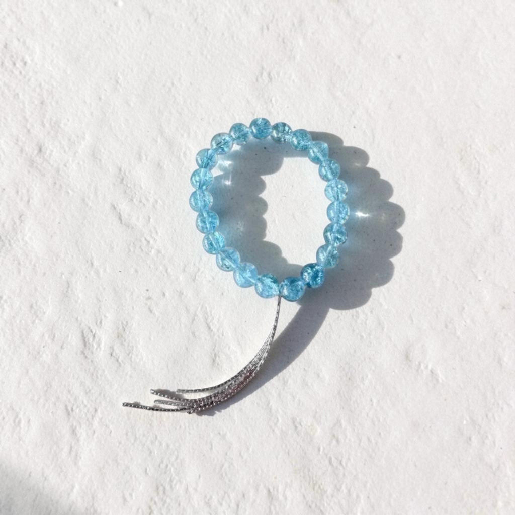 A light blue beaded stretch bracelet with a silver-toned metal tassel, photographed on a textured white background.
