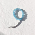 A light blue beaded stretch bracelet with a silver-toned metal tassel, photographed on a textured white background.