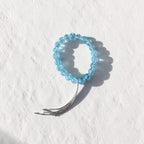 A light blue beaded stretch bracelet with a silver-toned metal tassel, photographed on a textured white background.