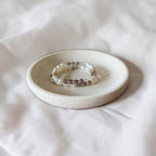 A beaded stretch bracelet with white pearls and brown smoky beads resting on a small round ceramic dish over a white fabric background.