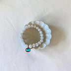 A beaded stretch bracelet with white round beads and a small turquoise-blue oval charm in a gold bezel setting, displayed on a small white ceramic dish against a textured off-white background.