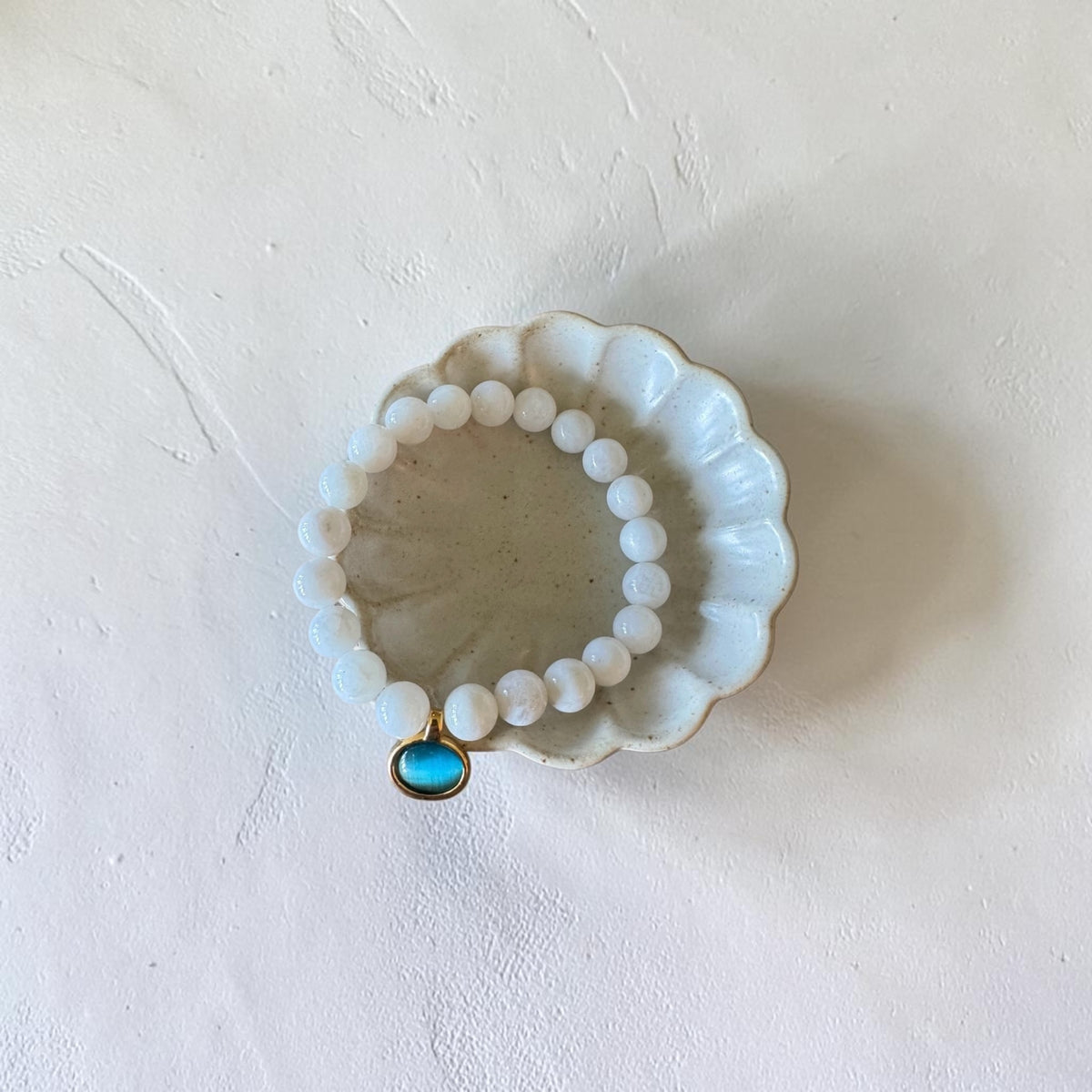 A beaded stretch bracelet with white round beads and a small turquoise-blue oval charm in a gold bezel setting, displayed on a small white ceramic dish against a textured off-white background.