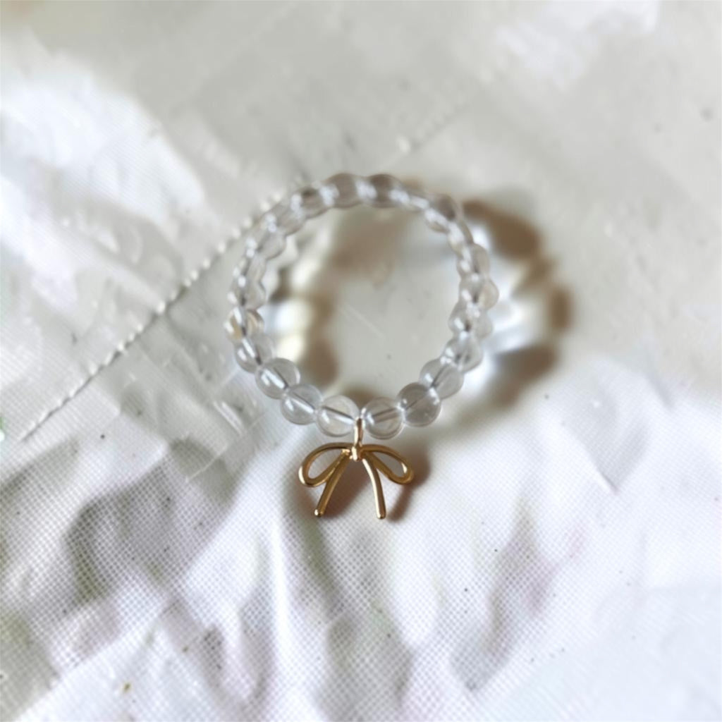 A beaded ring featuring clear, round glass beads and a small gold bow charm, resting on a textured white fabric background.